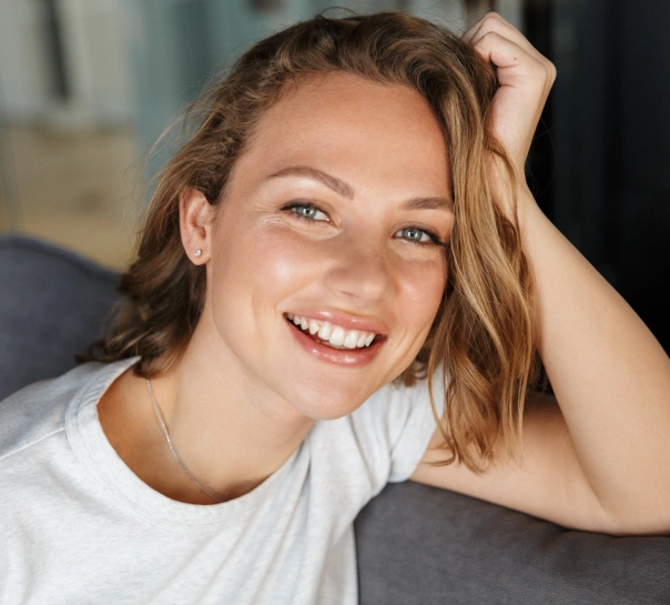 Young woman smiling at camera, hand in hair, wearing white shirt indoors.