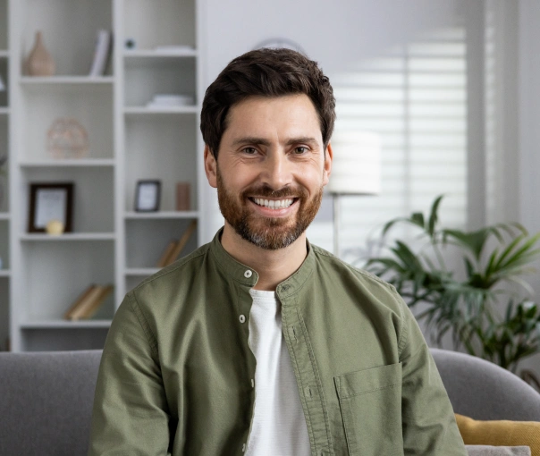 Smiling man in olive shirt sitting on couch in modern home office with shelving and plants.