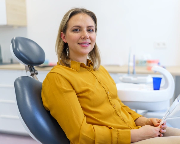 Female dentist in yellow shirt seated in dental office chair, smiling at camera.
