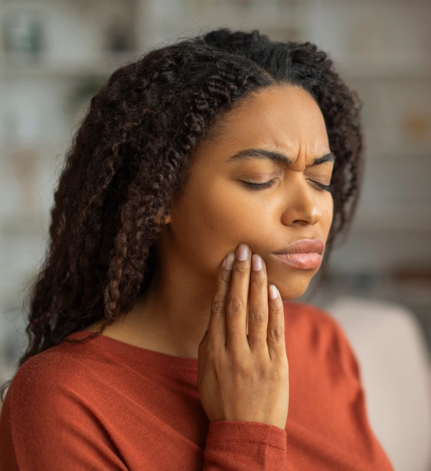 Woman in orange shirt experiencing tooth pain, holding jaw with hand indoors.