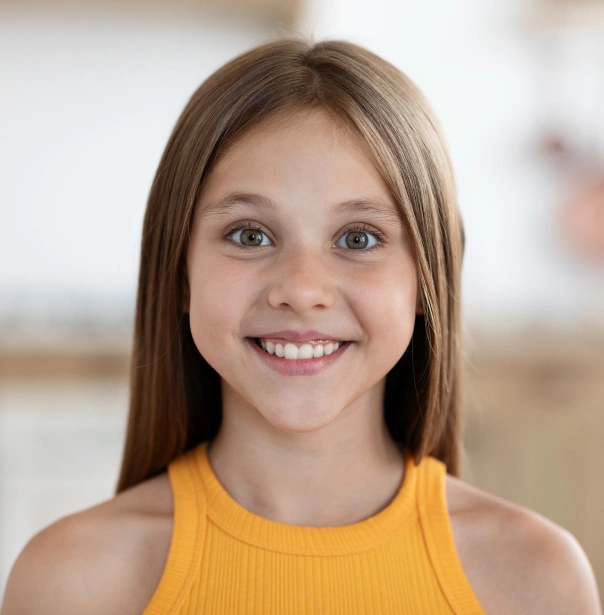 Young girl smiling at camera wearing yellow tank top, indoor blurred background