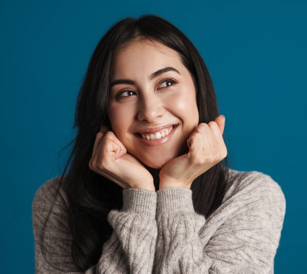 Woman smiling at camera with hands near face, blue background, professional portrait.