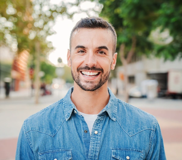 Professional headshot of smiling man in denim shirt, outdoor urban setting.