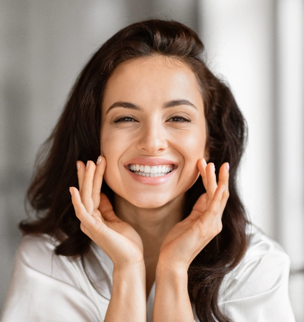 Smiling woman with dark hair wearing white shirt, hands near face, professional headshot in bright indoor setting.