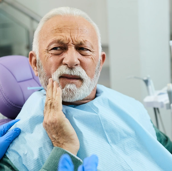 Senior man in dental bib touching cheek in discomfort at dental office visit.