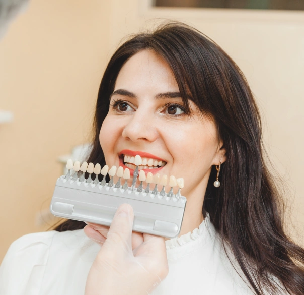 Woman holding dental shade guide near teeth, smiling in dental office setting.