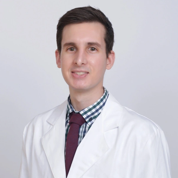Male dentist in white coat and checkered shirt smiling at camera against neutral background.