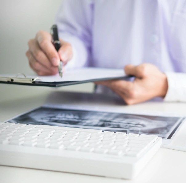 Dentist writing on clipboard at desk with laptop and dental X-ray visible.