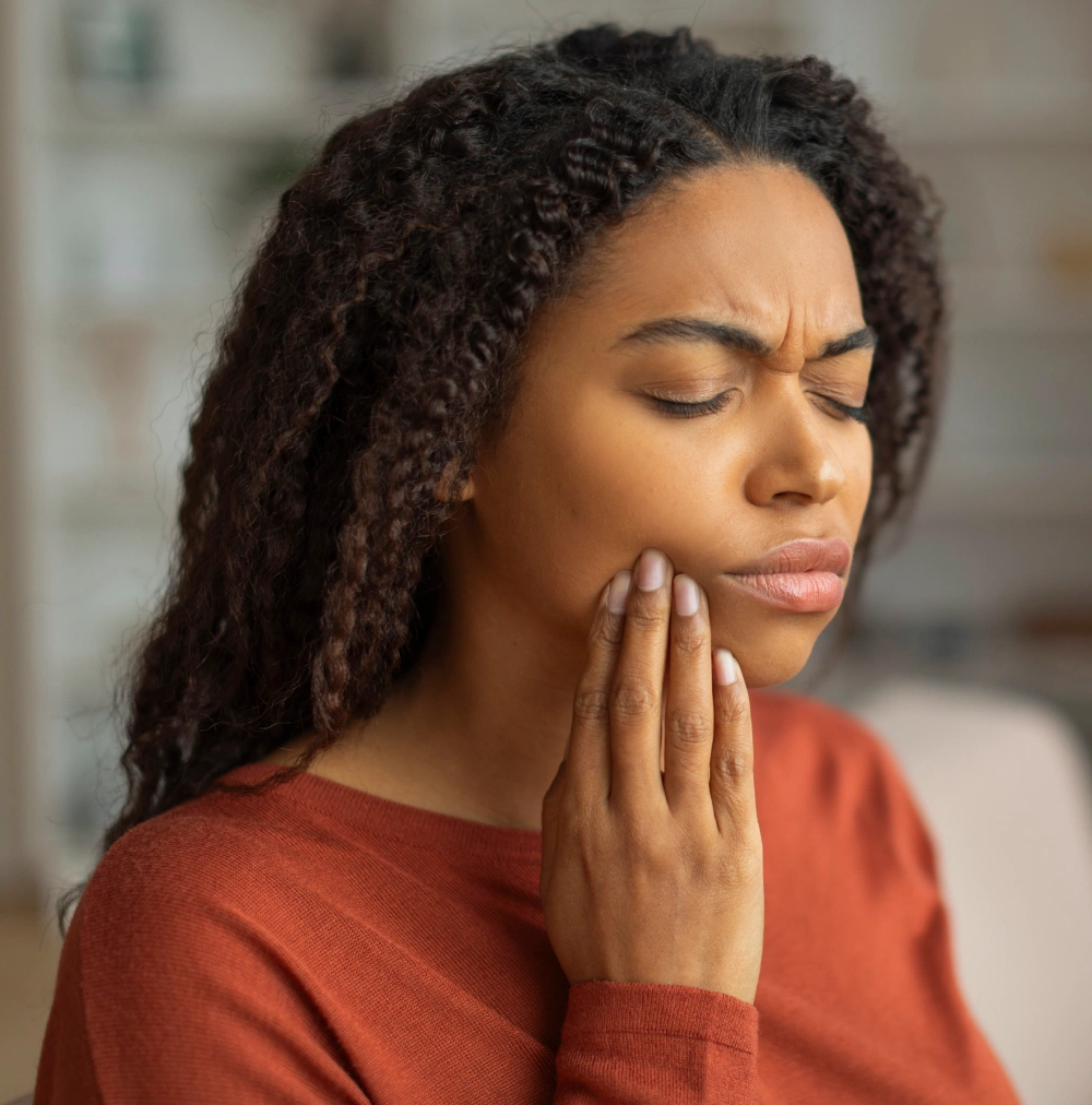 Woman in red shirt holding jaw, appearing to experience tooth pain or dental discomfort indoors.