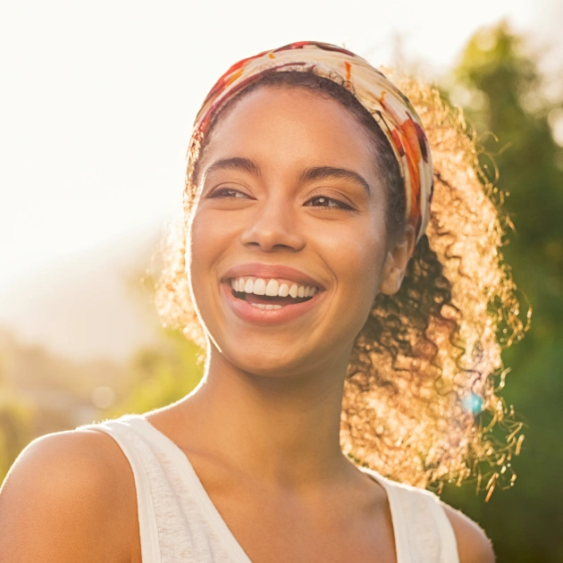 Smiling woman with curly hair wearing headband outdoors in golden sunlight.