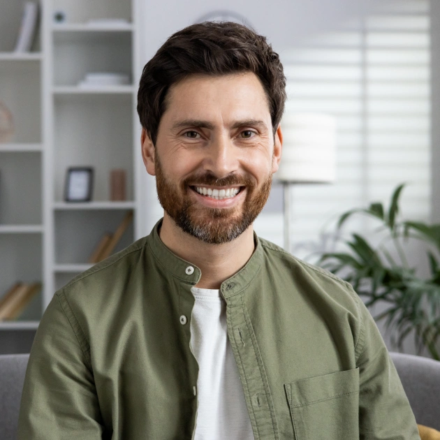 Smiling man in olive shirt seated indoors with shelving and plant in background.