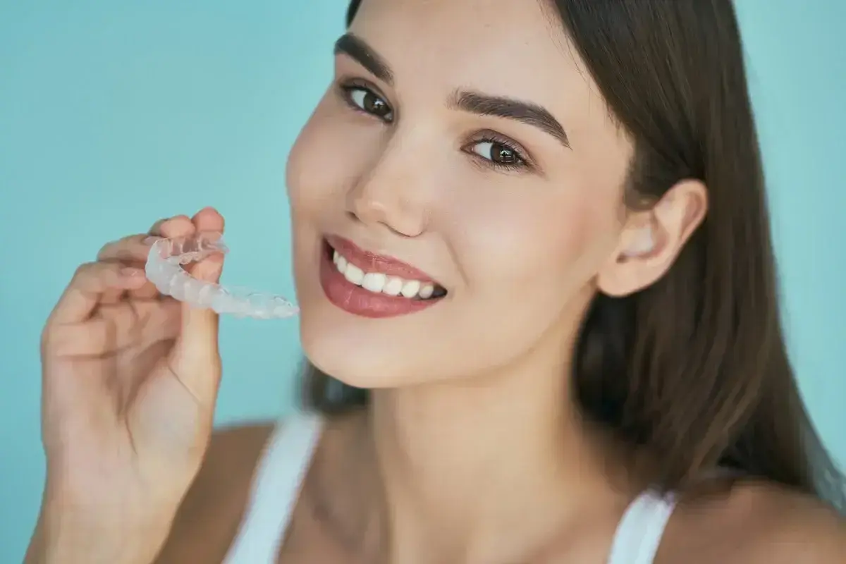 Woman holding clear Invisalign aligner, smiling against blue background.